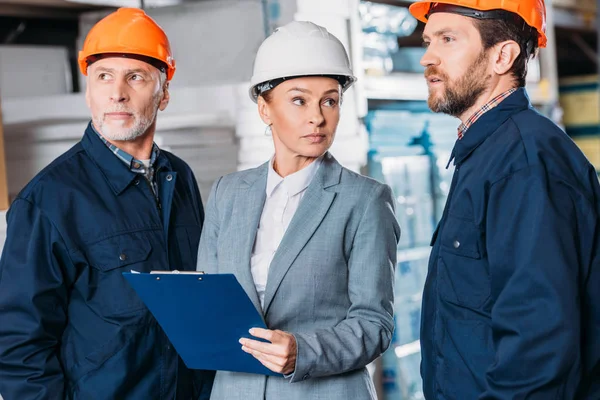 Male workers in helmets and female inspector with clipboard in warehouse — Stock Photo