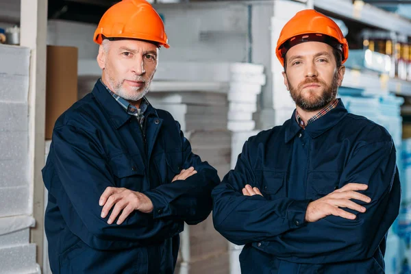 Trabalhadores do sexo masculino em capacetes posando com braços cruzados no armazém — Fotografia de Stock