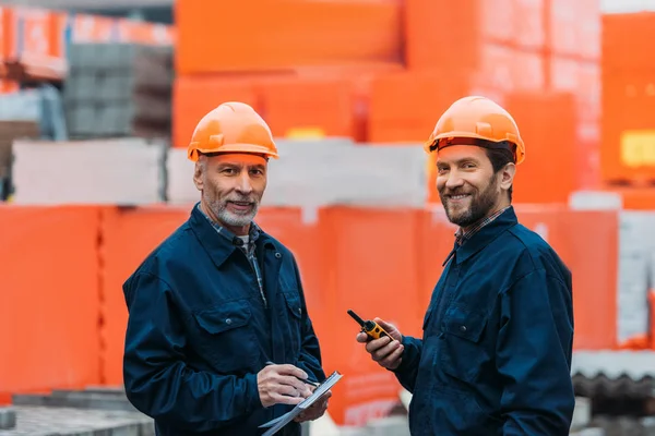 Deux constructeurs de casques travaillant avec talkie walkie et bloc-notes à l'extérieur sur la construction — Photo de stock