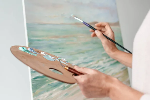 Cropped shot of artist holding palette and paintbrush while painting at easel in art studio — Stock Photo