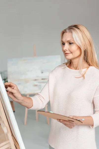 Beautiful mature female artist painting on easel in art studio — Stock Photo