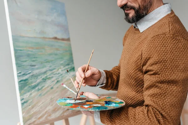 Cropped shot of bearded man holding palette and painting on easel at art class — Stock Photo