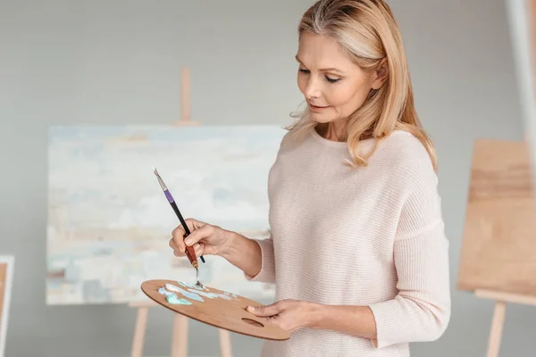 Mulher madura segurando paleta e pincel no estúdio de arte — Fotografia de Stock