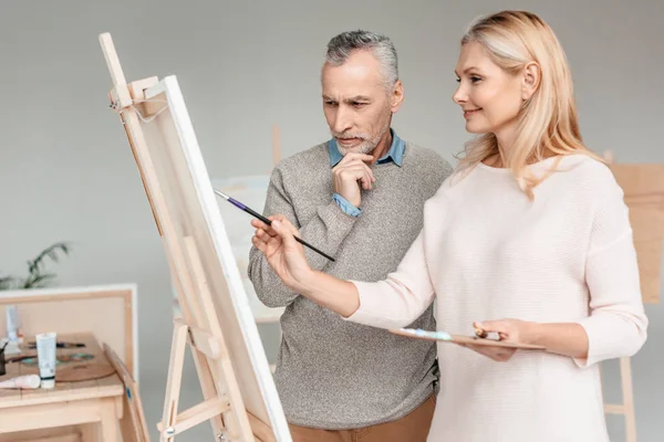 Senior homme regardant chevalet tout en souriant femme mature peinture sur elle pendant le cours d'art — Photo de stock