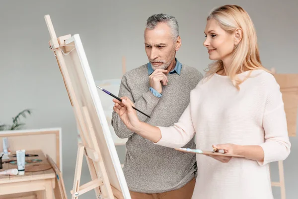 Uomo anziano guardando cavalletto sorridendo donna matura pittura su di esso durante la classe di arte — Foto stock