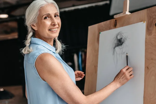 Schöne Seniorin lächelt in die Kamera, während sie im Kunstunterricht mit Bleistift auf Staffelei zeichnet — Stockfoto