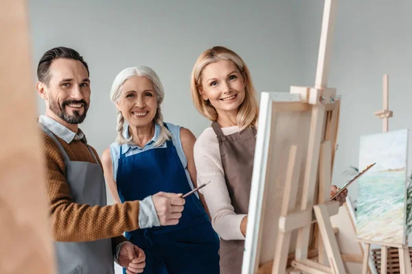 Allegri artisti maturi sorridenti alla macchina fotografica mentre dipingono insieme in studio d'arte — Foto stock