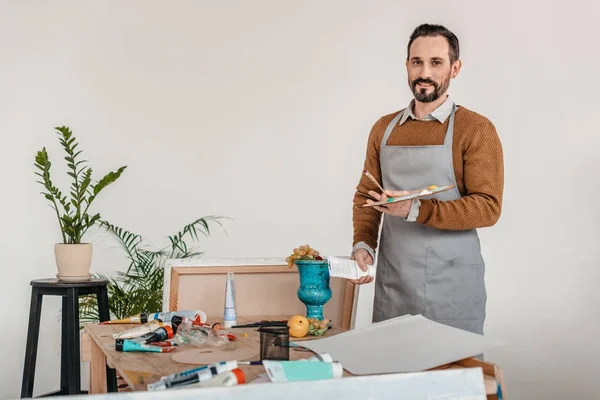 Handsome mature male artist in apron holding palette and smiling at camera — Stock Photo