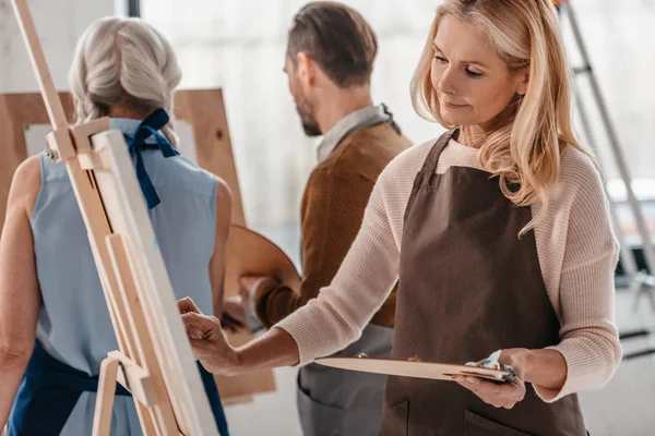 Hermosa mujer madura sosteniendo paleta y pintura en caballete durante la clase de arte para adultos - foto de stock