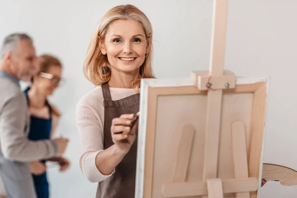 Красивая зрелая женщина улыбается перед камерой во время рисования на мольберте в художественном классе — стоковое фото