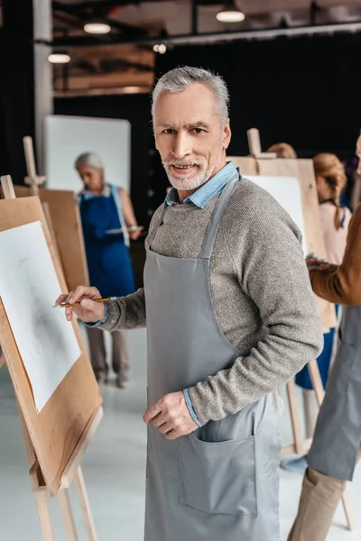Senior man smiling at camera while standing near easel at art class — Stock Photo