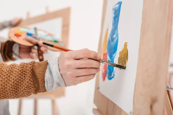 Recortado tiro de persona sosteniendo pincel y pintura en caballete - foto de stock