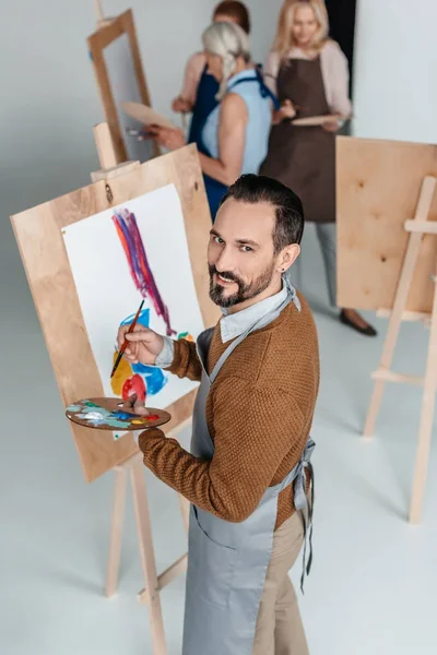Bonito maduro artista segurando paleta enquanto de pé perto cavalete e sorrindo para a câmera — Fotografia de Stock