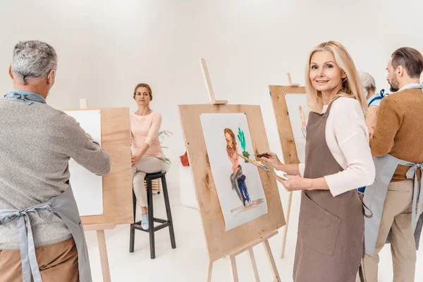 Femme mûre souriant à la caméra tout en peignant sur chevalet à la classe d'art — Photo de stock