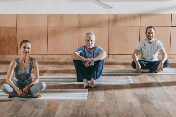 Инструктор-женщина со зрелыми мужчинами, сидящими на ковриках для йоги и улыбающимися в камеру — стоковое фото