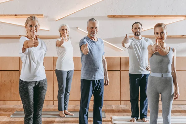 Personas mayores e instructor en ropa deportiva mostrando los pulgares hacia arriba y sonriendo a la cámara en el gimnasio - foto de stock