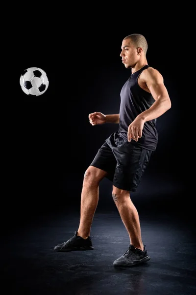 Handsome african american soccer player bouncing ball on leg on black — Stock Photo