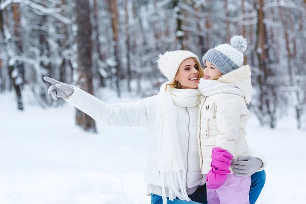 Улыбающаяся мать обнимает дочь и указывает на нее, стоя в зимнем парке — стоковое фото