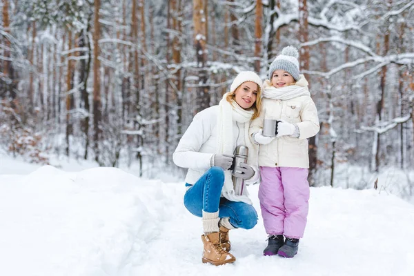 Familia con té caliente en tazas de termo mirando a la cámara en el bosque de invierno - foto de stock