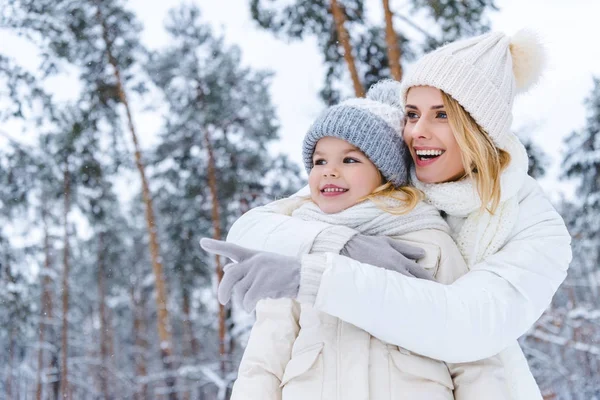 Улыбающаяся мать обнимает дочь и указывает на нее, стоя в зимнем парке — стоковое фото