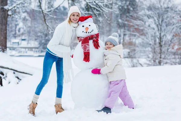 Счастливые мать и дочь, стоящие рядом со снеговиком в зимнем лесу — Stock Photo