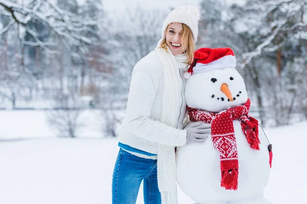 Porträt einer fröhlichen jungen Frau, die neben einem Schneemann steht und in die Kamera blickt — Stockfoto