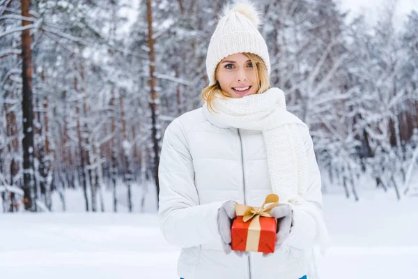 Beautiful young woman holding gift box and smiling at camera in winter park — Stock Photo