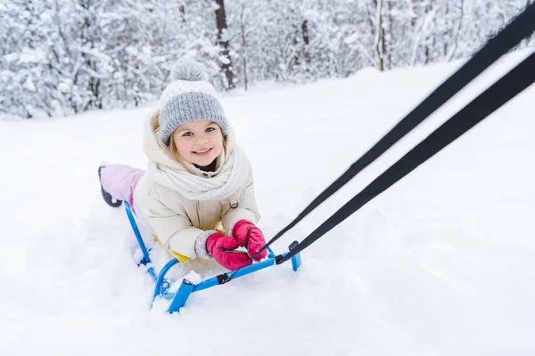 Lindo niño pequeño acostado en trineo y sonriendo a la cámara en el parque de invierno - foto de stock