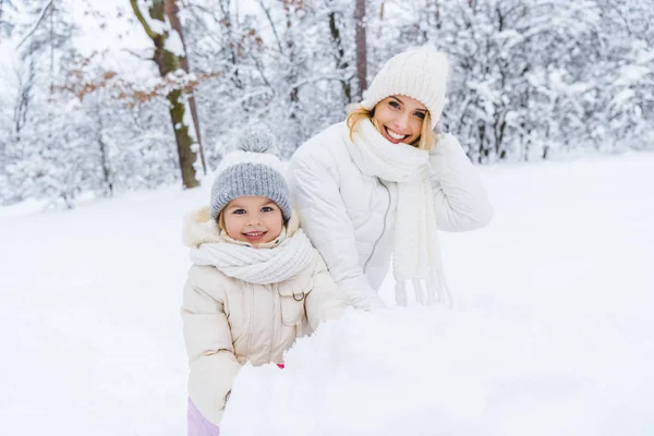 Schöne glückliche Mutter und Tochter lächeln in die Kamera, während sie Schneemann im Winterpark machen — Stockfoto