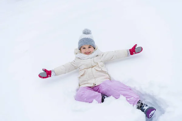 Adorable niño pequeño con los brazos abiertos tumbado en la nieve y sonriendo a la cámara - foto de stock