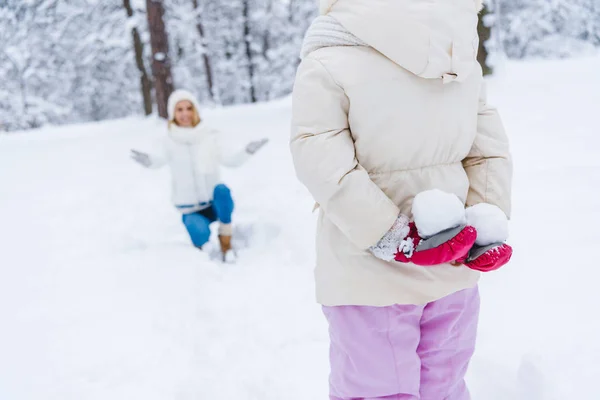 Recortado disparo de pequeña hija sosteniendo bolas de nieve y feliz madre arrodillado detrás en invierno parque - foto de stock