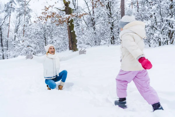 Feliz madre arrodillada con los brazos abiertos e hija pequeña caminando en la nieve en el parque de invierno - foto de stock