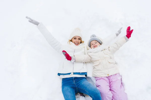 Feliz madre e hija con los brazos abiertos sonriendo a la cámara mientras yacen juntas en la nieve - foto de stock