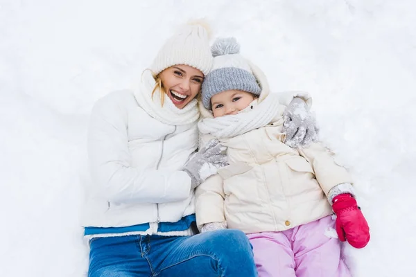 Mãe feliz e filha sorrindo para a câmera enquanto deitados juntos na neve — Fotografia de Stock