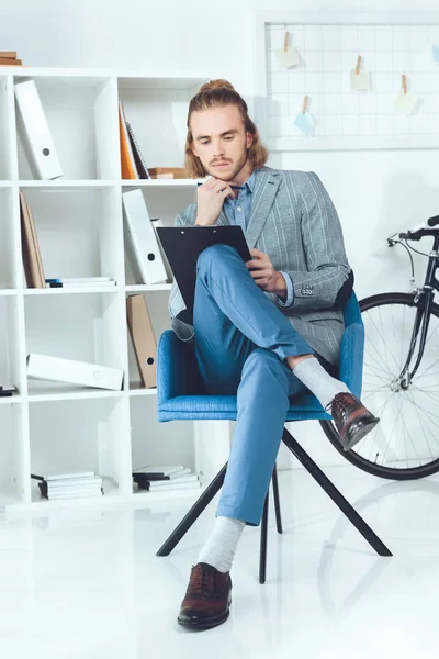 Pensive businessman looking at clipboard and sitting in chair — Stock Photo