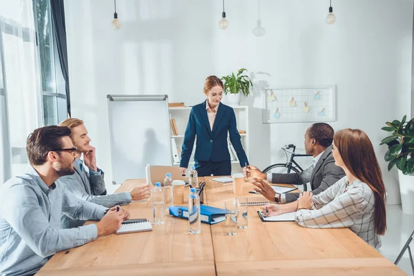 Mujer de negocios en traje formal de pie contra la mesa y hablando con el equipo en el espacio de oficina - foto de stock