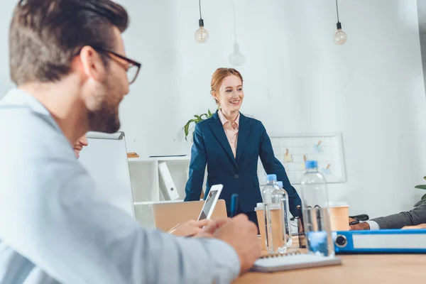 Mujer de negocios en traje formal de pie contra la mesa y hablando con el equipo en el espacio de oficina - foto de stock