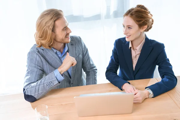 Empresarios sonriendo el uno al otro mientras se sientan en la mesa con el ordenador portátil - foto de stock