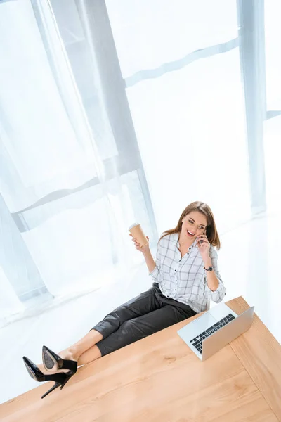 Mujer feliz en camisa sentada en la silla con las piernas en la mesa contra el ordenador portátil mientras habla por teléfono - foto de stock