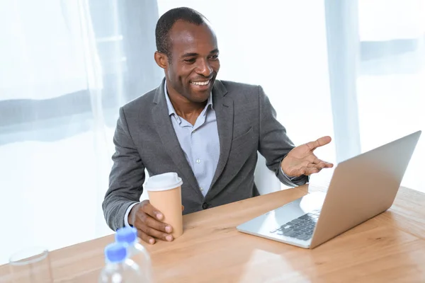 Hombre africano sonriente con taza de café sonriendo mientras mira en el portátil en la mesa - foto de stock