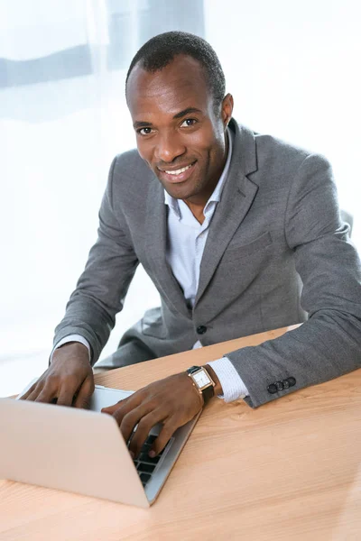 Улыбающийся африканский мужчина смотрит в камеру, используя ноутбук на столе — стоковое фото