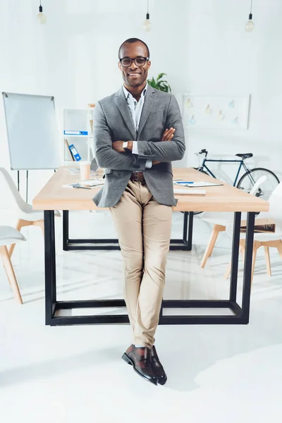 Усміхнений африканський чоловік спирається на стіл з обіймами, схрещеними в офісному просторі — стокове фото