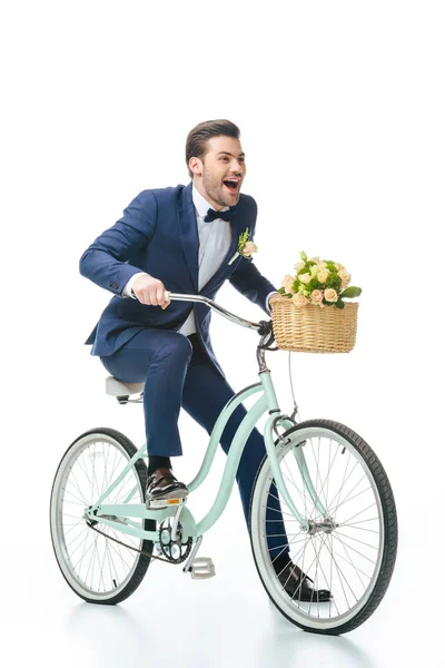 Happy groom in suit riding retro bicycle with wedding bouquet in basket isolated on white — Stock Photo