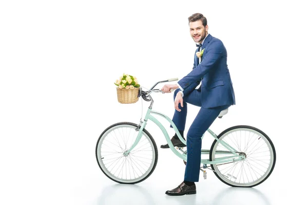 Groom in suit sitting on retro bicycle with wedding bouquet in basket isolated on white — Stock Photo