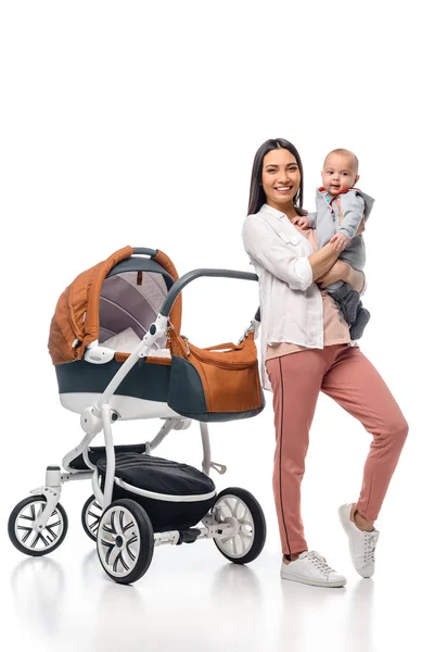 Madre feliz con bebé en las manos mirando a la cámara con carro de bebé detrás aislado en blanco - foto de stock