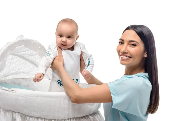 Side view of smiling mother taking baby out of crib isolated on white — Stock Photo