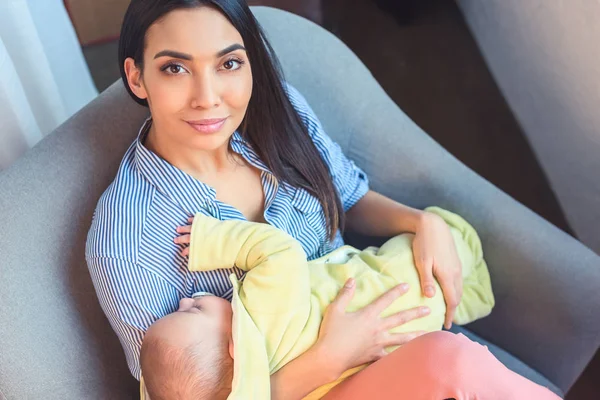 Visão de alto ângulo da mulher sorridente com o bebê nas mãos olhando para a câmera enquanto descansa na cadeira de braço — Fotografia de Stock