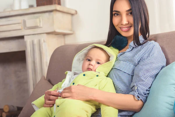Ritratto di madre sorridente con bimbo in mano appoggiato sul divano di casa — Foto stock