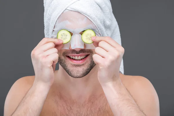 Lächelnder bärtiger Mann mit Kollagenmaske und Gurkenscheiben auf den Augen, isoliert auf grau — Stockfoto