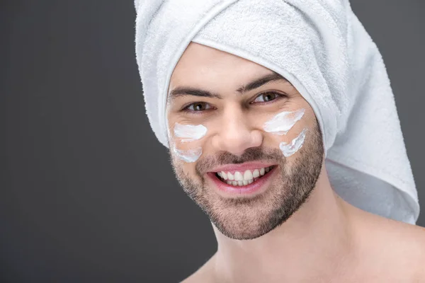 Smiling man in towel with cream on face, isolated on grey — Stock Photo